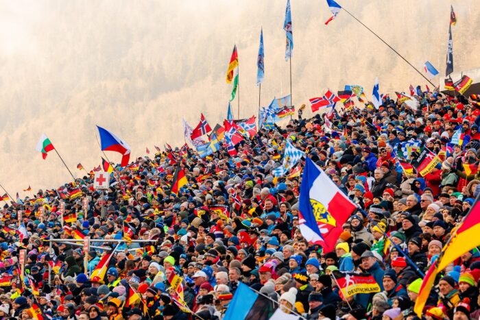 Audiodeskription-Biathlon-fuer-alle-erlebbar_gt2a6984_0 Gemini hat gesagt Fans mit vielen internationalen Flaggen auf der dicht besetzten Zuschauertribüne der Chiemgau Arena während eines Biathlon-Wettkampfs.