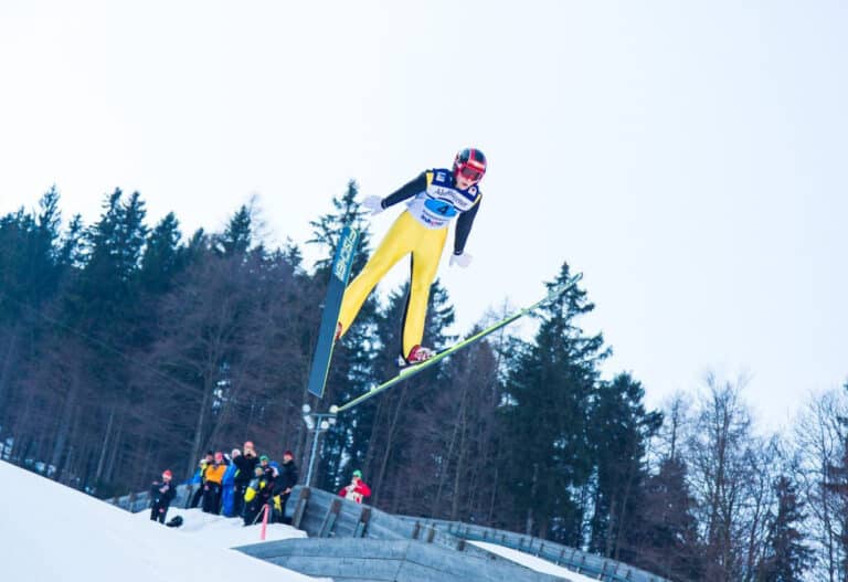 Skispringer im gelben Anzug im Flug über der Aufsprungbahn, Zuschauer am Rand der Schanze sichtbar.